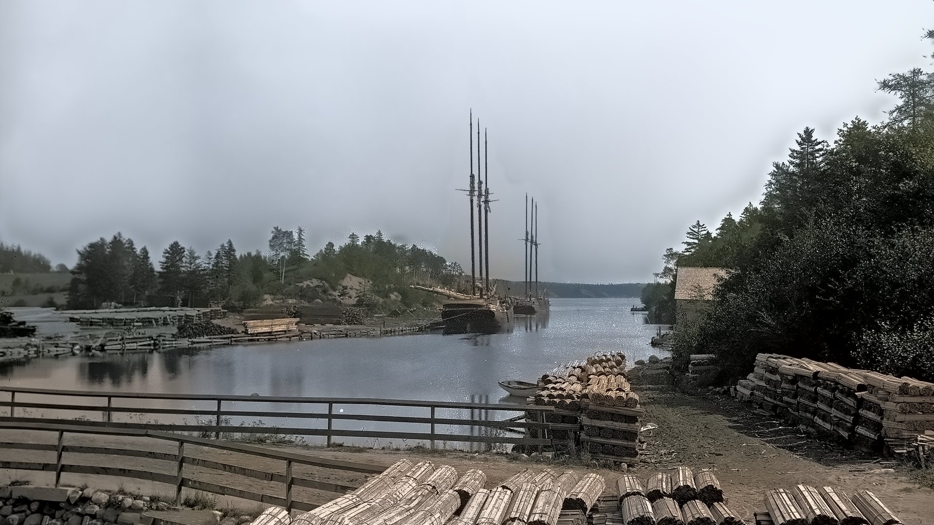 Chandler River in colour, a faithful recolour of the 1912 photograph: tall-masted schooners moored on the river, stacked timber on the bank and dark evergreens on the right.